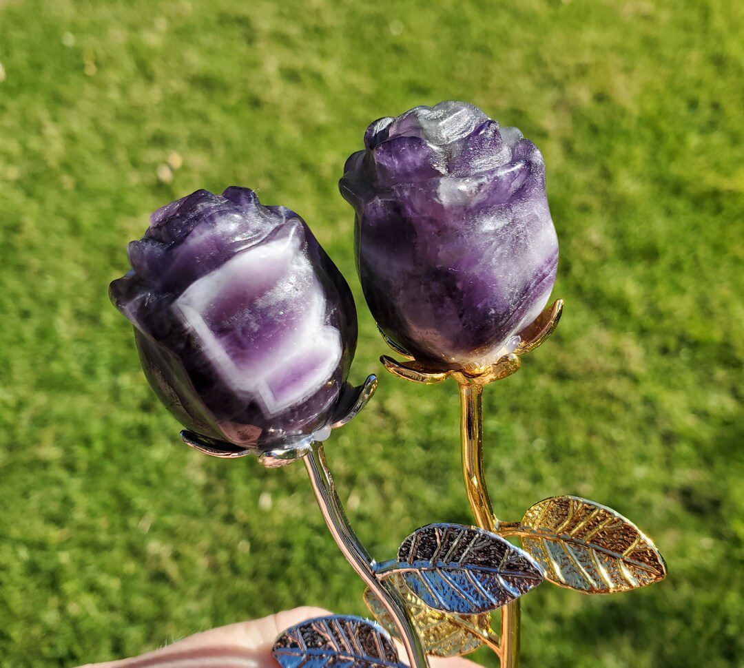 Hands supporting five purple and lavender crystal rosebuds on silver-colored stems and a small flared stand against a blurred green background
