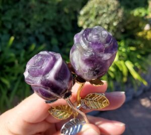 Hands supporting five purple and lavender crystal rosebuds on silver-colored stems and a small flared stand against a blurred green background