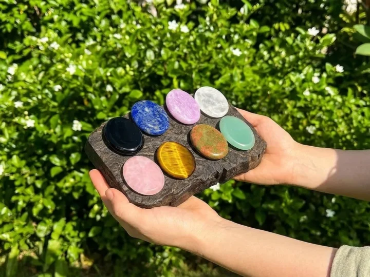 Ten polished oval worry stones in black, blue, pink, purple, clear, gold, and green, arranged on a wooden board held in two hands
